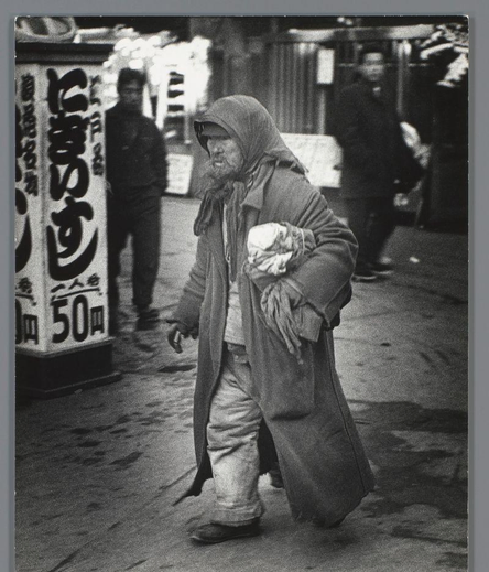 The image depicts a man in an urban street setting. He appears to be middle-aged, with weathered skin and a worn complexion suggesting exposure or hardship. The individual is wearing loose-fitting clothes; his attire consists of what seems like old-fashioned trousers tucked into boots at the lower end, which look tattered near their bottom hem. A long coat extends down past the knees, draped loosely over him without signs of wear typical for heavy usage.

The man carries a white bag in one arm and holds another item, possibly clothing or personal belongings wrapped in fabric, close to his body with his other hand. His expression is not clearly visible due to the angle but seems somber; he's looking off into space rather than directly at the camera. The street behind him has various signs that suggest commercial activity, including what looks like Japanese characters on a signboard next to which another man stands in the background.

The photograph captures this moment with high contrast and graininess characteristic of film photography from an earlier era, possibly mid-20th century given its style and quality. The context suggests it may be part of a larger collection or series documenting life at that time period, particularly focusing on individuals who might have been marginalized or less fortunate during the Cold War years in Japan as per historical records such as those referenced online.

Notably, there's no indica [...]