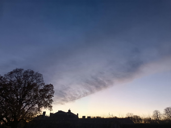 A froth of dark clouds smoulders across a fading blue sky as a descending sun simmers in over silhouettes of buildings among trees that line the horizon against a barely visible dark foreground.
