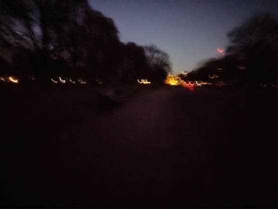 Moving camera captured colours of red through yellow to white hues line the horizon below a sky blue sunset sky amidst dark foliage of trees on both sides of tarmac of dark rouged highlights almost devoured by a ravenous approaching night.
