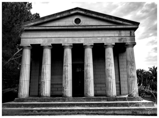 A low angle, black and white photograph of the imposing Seppelt Family Mausoleum at Seppeltsfield in the Barossa Valley. 

The Grecian style structure features six large Doric columns across the front, supporting a triangular pediment with a central round window (oculus).

Stone steps lead up to the portico. The dark shadows contrast sharply with the white stone and bright, cloudy sky.