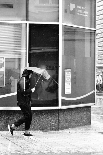 Das Foto zeigt eine Straßenszene in Schwarz-Weiß in der St. Pöltenter Innenstadt an einen regnerischen windigen Tag. Man sieht eine Person mit Regenschirm der durch den Wind verbogen ist.