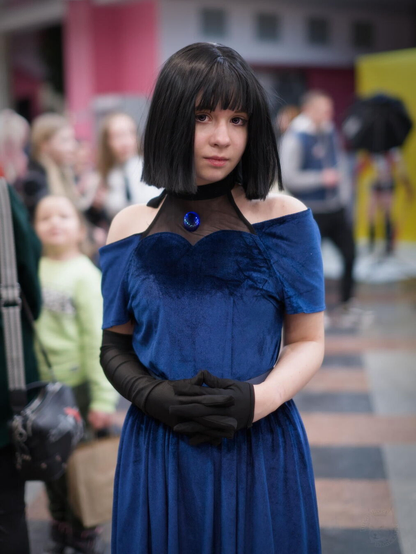 The image shows an individual with dark hair styled straight at shoulder length, wearing a blue dress with off-shoulder sleeves and adorned with a decorative brooch around the chest area. They are also dressed in black gloves that extend to their wrists. The person's hands are clasped together in front of them. In the background, there is an assortment of people who appear out of focus, suggesting this photo was taken at a public event or gathering where cosplaying might be present, as indicated by others' attire and props partially visible behind the individual in blue.