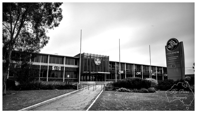 Black and white photograph of the long, two story administrative building for the former Holden (GMH) Manufacturing Operations, featuring a large circular Holden lion emblem above the entrance and a tall sign to the right displaying the Holden logo. A pathway leads up to the building entrance, flanked by landscaping.