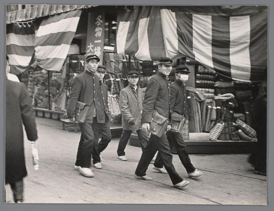 The image captures a moment on an urban street bustling with activity. A group of individuals, possibly students or young adults based on their attire and demeanor, are walking in unison across the pavement. The clothing style suggests a mid-20th-century time period; they wear dark coats and trousers, some carrying briefcases indicative of work or academic life.

They walk alongside market stalls draped with striped fabric banners that add texture to the scene, perhaps indicating a marketplace setting like Dotonbori in Osaka, Japan - known for its vibrant street culture. The focus is on their movement and camaraderie rather than individual identity; all faces are blurred for privacy, drawing attention away from personal features.

The grayscale tone of this black-and-white photo suggests it was taken before the widespread adoption of color photography or that a choice has been made to give it such an appearance post-processing. It evokes nostalgia while also offering insight into daily life and cultural settings of its time period in Japan.