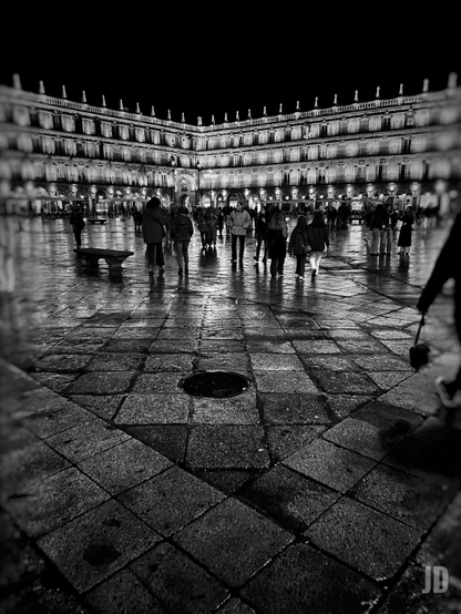 Fotografía en blanco y negro de una concurrida plaza de noche, con el suelo adoquinado y mojado que refleja las luces. En el primer plano, se observa una depresión circular oscura en el pavimento. Varias personas caminan por la plaza, algunas difuminadas por el movimiento. Al fondo, un gran edificio de arquitectura clásica, formando una esquina y con múltiples pisos y ventanas, está brillantemente iluminado contra el cielo nocturno