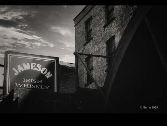 Black and white photo of a rustic stone building with a large Jameson Irish Whiskey sign. The sky is partially cloudy, creating a moody atmosphere.