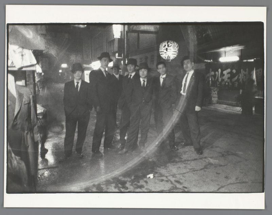 The image depicts a group of men standing on an urban street at night, with the lighting creating high contrast and visible shadows. They are dressed in suits and hats indicative of mid-20th-century fashion, suggesting a historical setting, likely from Japan given the Asian script lantern hanging above them. The scene appears to be taken candidly without posed postures or expressions, capturing an authentic moment rather than a staged portrait.

There is no visible indication of age within this group due to their attire and hairstyles being consistent with mid-century men's fashion trends. No particular individual stands out as the focus; instead, they all seem part of a collective unit. The environment around them includes what looks like a street in Japan at night time-lit by artificial sources such as lamps or car headlights.

The photograph is black-and-white, adding to its vintage feel and potentially suggesting it was taken with older photographic equipment which captured grayscale images before color film became commonplace for everyday use among amateurs.