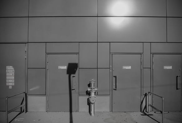 Das Foto zeigt einen Betriebsbereich einer U-Bahnstation in Wien in Schwarz-Weiß. Im Vordergrund siehst du drei große, graue Türen mit Beschilderungen wie "Betriebsraum" und dazugehörigen Raumnummern. Links neben den Türen befindet sich Schilder mit Sicherheitshinweisen.
Vor den Türen steht mittig ein Hydrant, und es gibt zwei Metallgeländer, die vermutlich als Barriere oder Absperrung dienen. Die Beleuchtung wirkt durch den Kontrast des Sonnenlichts und dem Schatten einer Straßenlaterne besonders.