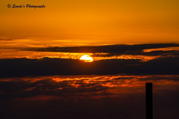 "The sun rises behind a mass of dark, heavy clouds, its light breaking through in molten streaks of orange, red, and gold. The sky itself becomes a fiery canvas, where shadow and brilliance wrestle for dominance. The clouds glow at their edges like embers, while their centers remain brooding and deep.

In the lower right, a vertical wood pile juts out of the ocean, silhouetted against the blaze of morning. Weathered and stark, it stands like a sentinel offshore, a quiet witness to the day’s first light. Its presence anchors the scene, reminding us of the human touch against the vastness of nature.

There is no shoreline visible—only sky, cloud, and the upright silhouette rising from the water. The atmosphere feels charged yet still, as if the Gulf itself is holding its breath while the sun forces its way into morning. It is a moment of reckoning between night and day, shadow and flame.

The image is signed “© Swede’s Photographs” in the top left corner, marking this sunrise as a sovereign act of witness." - Microsoft Copilot with edits