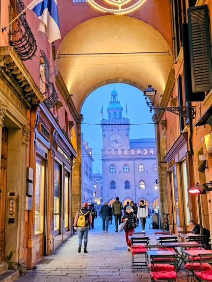 A warm-toned portico in Bologna opens onto a cobblestone street at dusk, framing a view of the clock tower on Piazza Maggiore against a blue evening sky. People in winter clothing walk beneath the arch toward the square, passing glowing shop windows on the left, while empty café tables with red cushions sit to the right. The soft lights under the portico and the historic architecture create a welcoming atmosphere.