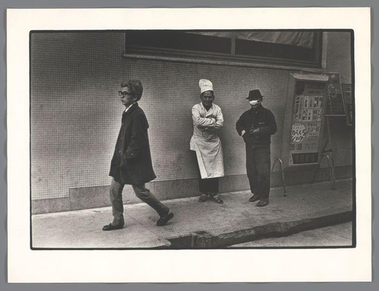 The image is a black and white photograph depicting three individuals standing on the sidewalk outside of what appears to be an establishment with glass windows. The person on the left, dressed in dark clothing including a coat and hat, is walking away from the camera towards the right side of the frame. This individual has their head turned slightly toward us as they move.

In the center stands another person wearing a white apron over casual attire, arms folded across the chest with an upright posture against the wall. To the right, there's someone in dark clothing including trousers and shoes, standing still with hands behind their back looking towards the camera.

Behind these individuals is a signboard displaying various texts and graphics that seem to be advertisements or information for potential customers inside the establishment they are outside of. The sidewalk appears aged with visible wear marks suggesting an urban setting possibly from several decades ago based on fashion styles and architecture design elements present in the image.

The context provided indicates this photograph was taken by Ed van der Elsken, a Dutch photographer known for his street photography work capturing moments of everyday life in various cities around the world. The specific location referenced is Dotombori in Osaka, Japan during 1960 which gives us insight into both the geographical and historical setting of the photo.