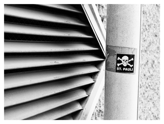 En liggande svartvit bild på ett klistermärke på ett stuprör. Märket har en korslagd döskalle med texten ”ST. PAULI”. Till vänster ett luftintag bakom ett galler.