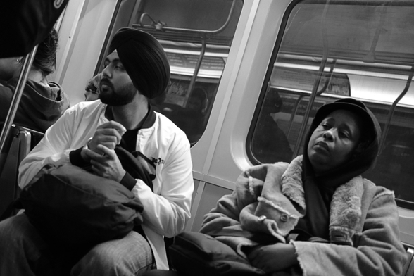 The image is tilted, the left side higher, as if taken on the fly. In perpendicular seats sit a bearded Sikh man in a turban and black woman. He is looking at something beyond the left edge of the frame. Her eyes are closed, her head tilted toward the right of the frame. Behind them, the lights of the subway car are reflected in the glass of the windows.