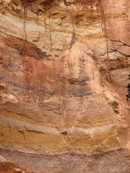 A closeup of an eroded section of earth displaying beautiful layers of colors, including shades of pink, rust, and ochre yellow and lavender. It looks like an abstract expressionist painting with horizontal layers of color and rust color drips traveling down throughout the frame. 