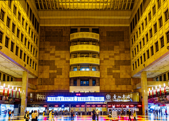 Atrium-like ticketing hall with ticket counters, people, neon signs, interior balconies, and “Taipei Main Station” lettering in Hanzi and English.