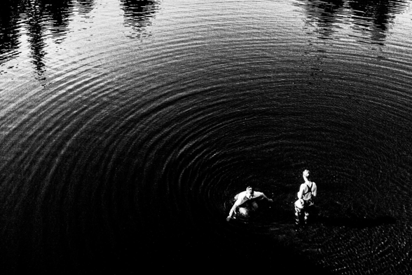 Black and white photo of two people wading in water with concentric ripples around them.