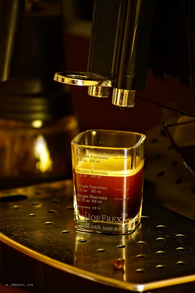 Unter dem Ausguss eines Kaffeevollautomaten steht ein Espresso-Glas. Das Glas ist mit Gravuren versehen, die anzeigen, wie groß ein Espresso oder ein Ristretto ist. Oder eben die doppelte Version der beiden... 
Das Glas ist voll, der Kaffee schäumt noch nach. Und das Glas steht auf einem Lochblech. 