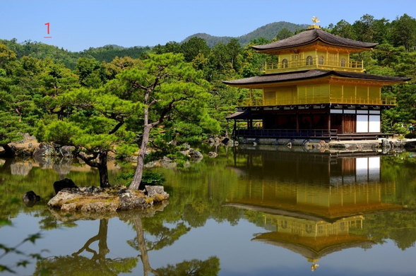 Photo couleur d'un temple recouvert d'or qui se reflète dans un étang. À gauche du temple qui est à droite sur la photo, un îlot où poussent des pins En arrière-plan des reliefs boisés. Au-dessus une partie du ciel qui est bleu.