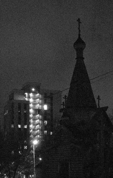 In the foreground is the silhouette of a wooden Orthodox church. In the background is a high-rise modern building with glowing balconies.