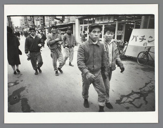 The image is a black and white photograph depicting a group of individuals walking down an urban street. The main focus is on three men in the foreground, with one prominently featured man wearing a jacket and pants that appear worn or dirty. He has short hair and looks directly at the camera with a neutral expression.

Behind him are two other men dressed similarly; their attire suggests they may be working-class individuals, possibly involved in manual labor given the state of their clothing. All three have boots on, which is consistent across all visible footwear among them.

In the background, there's another group of people walking away from the camera. They appear to be a mix of men and women wearing casual street clothes typical for that era. One woman holds an umbrella with Japanese writing on it, suggesting this scene might take place in Japan or involves Japanese-speaking individuals.

On the right side of the image is a bicycle leaning against what appears to be a shop's exterior wall. The storefront has signage with more Japanese characters, indicating commercial activity along the street.

The setting suggests a busy urban area with various shops and pedestrians moving about their day. The ground shows some wear or damage in places, which adds texture and context to the environment depicted.