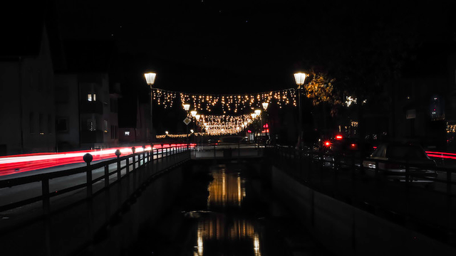 Die Hauptstrasse durch unseren Ort bei Dunkelheit fotografiert. Durch die längerer Belichtungszeit sieht man von den Autos auf der linken Seite nur die Lichtspuren. In der Mittel läuft der Dorfbach. Rechts und links davon sien Strassenlaternen in einigen Abständen positioniert. Zwischen diesen Strassenlampen sind Lichterketten als Adventsschmuck montiert. Die Lichter spiegeln sich im Dorfbach.