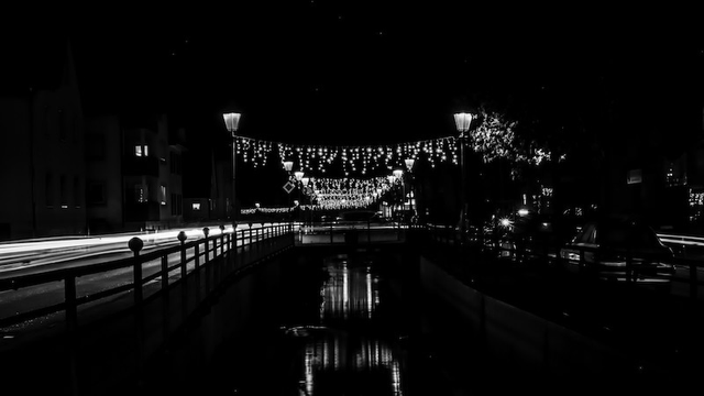 Die Hauptstrasse durch unseren Ort bei Dunkelheit fotografiert. Durch die längerer Belichtungszeit sieht man von den Autos auf der linken Seite nur die Lichtspuren. In der Mittel läuft der Dorfbach. Rechts und links davon sien Strassenlaternen in einigen Abständen positioniert. Zwischen diesen Strassenlampen sind Lichterketten als Adventsschmuck montiert. Die Lichter spiegeln sich im Dorfbach. Dieses Foto ist in Schwarzweiß.