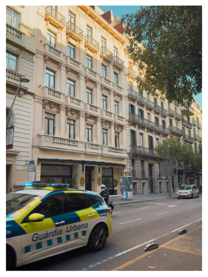 A city street in Barcelona with late-afternoon sunlight on the façades of elegant, historic apartment buildings. In the foreground, a “Guàrdia Urbana” police car with blue lights drives past, while a police officer on a motorcycle waits nearby. A few cars are further down the road, and tall trees partially frame the scene on the right.