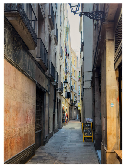 A narrow pedestrian alley in Barcelona, lined with tall, closely packed buildings in warm earth tones. Small balconies with iron railings jut out above the street, while decorative lamps hang from ornate brackets. Sunlight filters in from the far end, where a few shops display colorful items. A yellow sandwich board advertising a tour stands on the right side of the alley, adding a touch of brightness to the shaded passageway.
