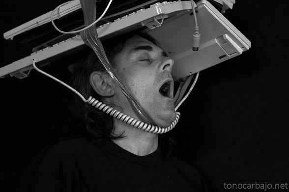 retrato en blanco y negro. Cabeza de un hombre con la boca abierta y ojos cerrados, sobre su cabeza atados con cinta de embalar varios teclados de ordenador