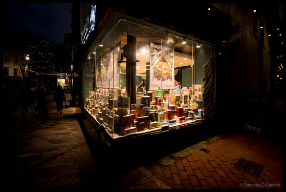 A night-time photograph of Dubray bookshop storefront in Cork with illuminated window display featuring books arranged artfully under a sign reading "BOOKS FOR CHRISTMAS", warm interior lighting highlighting multiple book covers in red, green and gold tones, dark street with pedestrians and decorative festive lights visible in the background.