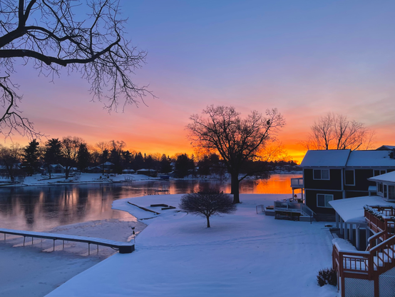 Photograph of an orange and yellow sunrise behind a narrow frozen lake with snow, houses, and bare trees on either shore.
