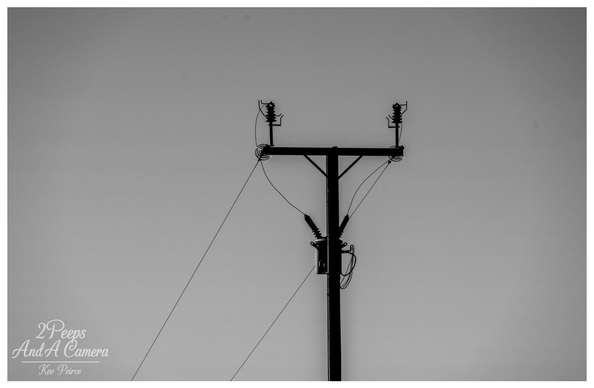 A high contrast, black and white photograph focusing on the top of a utility pole and its electrical lines, silhouetted against a smooth, bright gray sky.

The stark, geometric shapes of the insulators and transformer create a strong visual cross shape in the center of the frame, giving the image a graphic, minimalist feel. Photo by Kev Peirce.