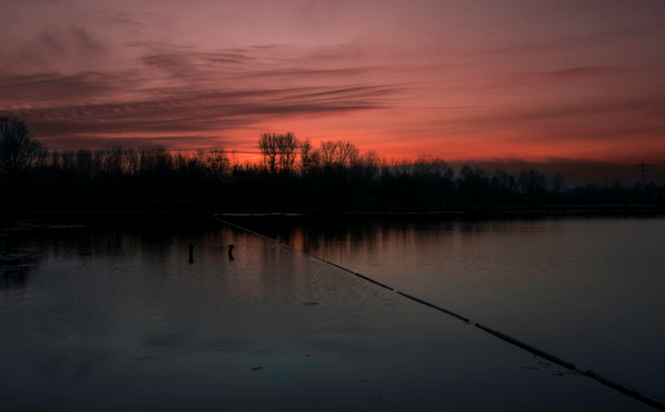 Ein rötlicher Himmel, nachdem die Sonne untergegangen ist. Davor ist ein Wald und davor ein See. Am Himmel sind kleine Schleierwolken zu sehen.