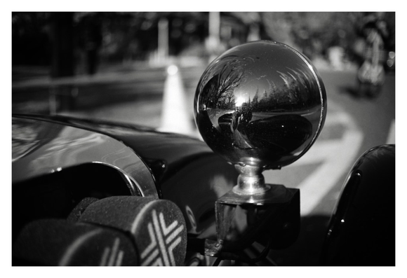 Black and white close-up of the headlight of a vintage hot rod. Rows of threes can just be made out reflected in the chrome.