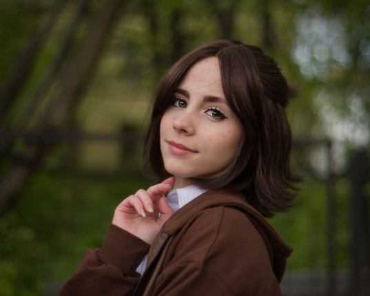 The image is a portrait of a young woman, likely taken outdoors in natural light. It has a slightly soft and dreamy feel due to the blurred background. She has a fair complexion with warm skin tone. Her hair is a medium-brown color and styled in a slightly messy, layered bob. It appears to be slightly wavy. She is looking slightly off to the side, with a gentle and contemplative expression. Her lips are subtly curved, giving a feeling of warmth and thoughtfulness. She's wearing a brown hoodie with a white shirt underneath. The hoodie is pulled up slightly, and a visible ribbed collar suggests it's a casual outfit. She's holding one hand up to her chin, with her fingers lightly touching her cheek - a gesture of reflection or contemplation. The background is a blurred expanse of green, suggesting trees. The depth of field effect makes the background soft and muted, keeping the focus firmly on the woman.