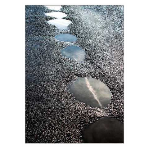 On an asphalt road, the wheels of heavy cars have created depressions, these are circular depressions in which water has stagnated after rain and in which the sky is reflected.