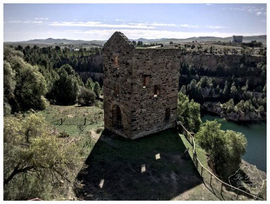 Ruins of the Morphett's Engine House, a historical stone building standing on the edge of the deep Burra Mine open cut, overlooking the vivid turquoise water below.

The scene is set under a bright blue sky, surrounded by green hills and trees.