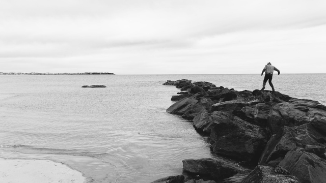 A man looks tiny in the context of a rock jetty he is walking across made of very dark rock in this black and white photo