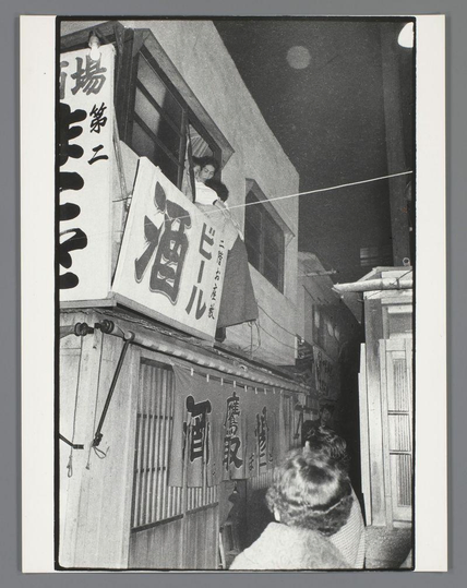The image is a black and white photograph depicting an urban alleyway at night. There's prominent signage in Japanese, with one large sign reading "酒" which translates to 'alcohol' or more specifically 'beer'. The smaller signs have different symbols, possibly representing businesses like bars or restaurants along the street.

A woman appears on top of a structure within this alley; she is partially obscured by an overhanging part of the building. She seems to be in mid-conversation with someone not visible in the frame and is wearing what looks to be traditional Japanese attire, including perhaps a kimono-like garment draped across her shoulders. The focus shifts toward the back where another individual's head enters from below.

The setting appears somewhat gritty or lived-in, given the wooden structures of buildings and hanging signs that suggest an urban nightlife scene in Japan.