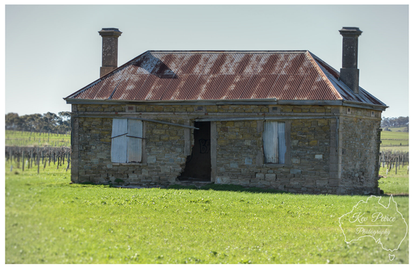 Stone ruins of an old, abandoned farmhouse with a rusted corrugated iron roof, two tall brick chimneys, and boarded up windows, set in a vibrant green field.  The stone walls are partially crumbled around the front doorway, revealing the darkness inside. A vineyard and trees are visible in the background under a pale blue sky. Signed Kev Peirce.