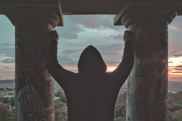 Guy with a hoodie (me) gazing at the sunset. Next to me are architectural poles of an abandoned building, and my hands are hanging between them. Colors are hued with teal and orange (mostly on sunset).