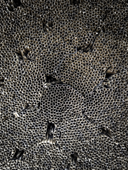 A huge pile of bundles of reed. The surface is dominated by the circular cross-sections of countless tubes packed tightly together. The repetitive texture resembles a honeycomb or the cells of an organism when viewed from a distance.

The color palette is monochromatic or near-monochromatic, featuring shades of grey, beige, and brown. If you look more closely, you can see the metal wire used to make the bundles as black and white, dotted lines.