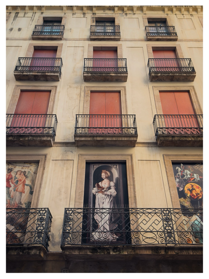 Façade of an old building in Barcelona with several small balconies and closed red shutters. On the ground-floor level, large framed images imitate windows, including a central depiction of an angel holding a chalice, flanked by two other classical-style artworks. The building’s pale, weathered exterior and ornate black iron railings give it a historic, slightly aged appearance.