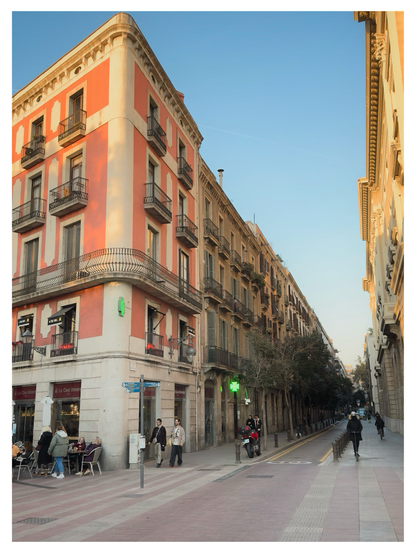 Street corner in Barcelona with a tall, salmon-and-white apartment building featuring numerous small balconies. A café terrace with seated patrons occupies the lower left, while pedestrians walk along the wide, sunlit street. On the right, a tree-lined lane stretches into the distance, flanked by historic buildings under a clear blue sky.
