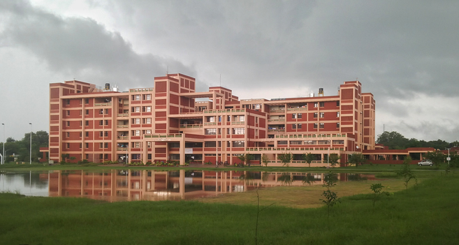 Red and white multi-story building reflected in a calm pond, set against a cloudy sky and surrounded by lush greenery.