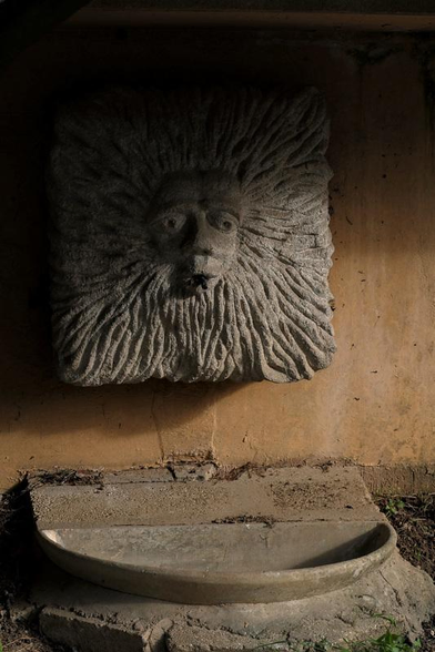 Sculpture d’une tête de Lion mais aplatie dans une dalle de pierre carrée. Un réceptacle est dessous, sûrement une fontaine en temps normal