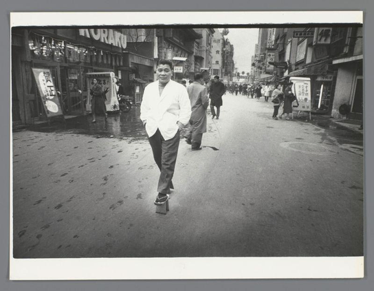 A black and white photograph capturing a moment on an urban street in Japan. The focus is on an individual dressed in traditional Japanese attire, specifically a kimono with an open front revealing trousers underneath. This person stands centrally, walking away from the camera towards a more populated area of shops and pedestrians lining both sides of the street.

The scene depicts everyday life, possibly in Osaka as indicated by text at top left that reads "KURA...," suggesting a storefront or signboard common to such urban environments. The streetscape is lively with numerous figures moving about; some are closer to the camera while others farther away near various commercial establishments displaying signs and banners.

Notable features include wet ground, indicating recent rain, which reflects light from overhead sources, possibly street lamps or shop lights. There's a visible contrast between those in traditional clothing like the central figure and more modern attire of passersby such as coats and trousers, highlighting cultural diversity within this urban setting.