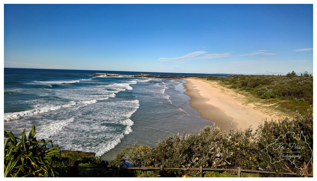 A sweeping, elevated view of a sandy beach on the Northern Rivers coast of New South Wales (NSW), south of Byron Bay.   Powerful white waves roll in from the deep blue ocean, meeting the golden sand. Coastal scrub and a wooden fence line the foreground, with a distant rock breakwater visible.