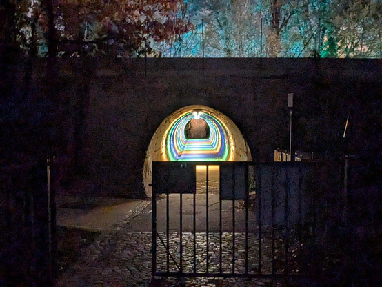 Rainbow lights inside a small walkable tunnel, gated. It's night time.