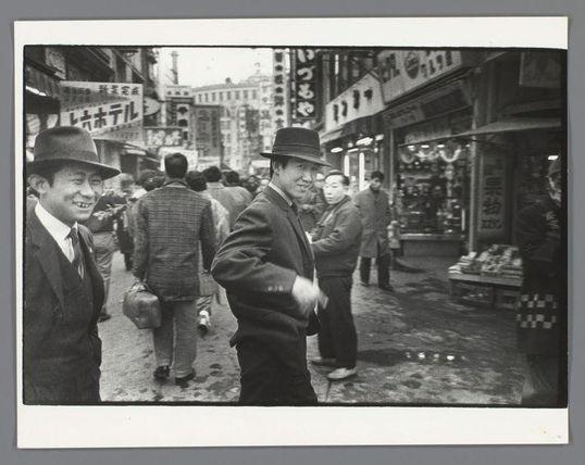 The image depicts a bustling street scene in what appears to be an urban Asian setting. The photograph captures several individuals walking or standing on the sidewalk, with varying expressions and postures that suggest movement and daily life activity.

In the foreground, two men are prominently featured: one wearing a dark suit and hat is smiling at the camera, exuding friendliness; the other, dressed in casual attire including a striped shirt and jacket, looks away from the lens. Behind them, there's another individual with a more serious expression walking towards the viewer.

The street behind these figures reveals an array of storefronts adorned with Japanese characters on signs, indicating that this may be Kamagasaki, Osaka—a known district for yakuza activities during van der Elsken’s time. The shops display various goods and merchandise, suggesting a lively marketplace atmosphere.

The overall composition of the photograph conveys a candid glimpse into street life in Japan at the beginning of the 1960s, with elements that hint at the socio-economic context as well as cultural aspects of urban living during this period.
